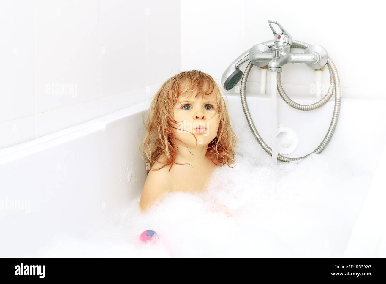 Serious blue-eyed toddler in the white bath. Infant washing and bathing. Hygiene and health care. Lips folded tube, eyebrows furrowed, looking up Stock Photo