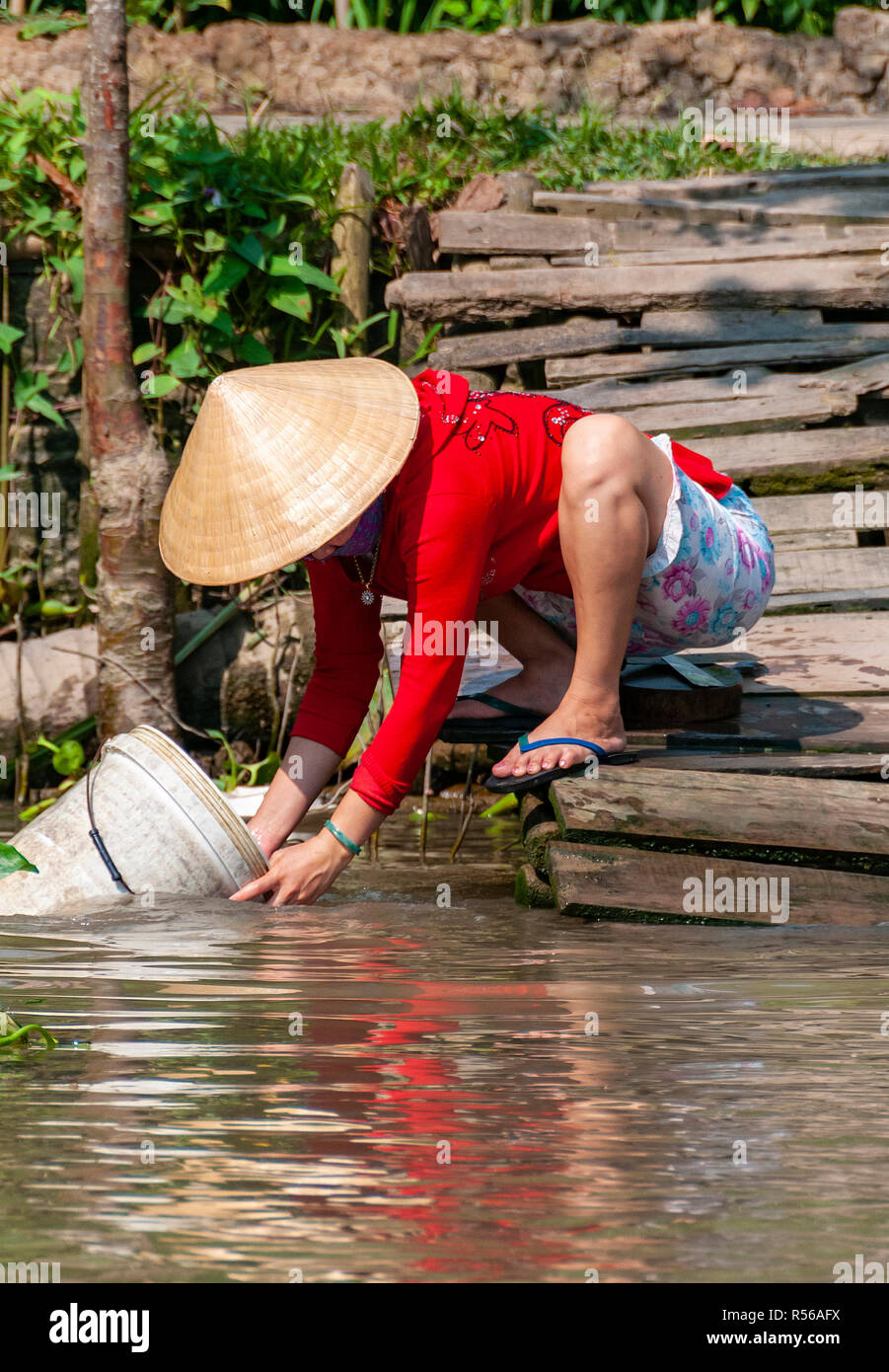 Young woman wearing bright red top crouching by the water's edge to wash out a bucket in river at Can Tho Province, Vietnam Stock Photo