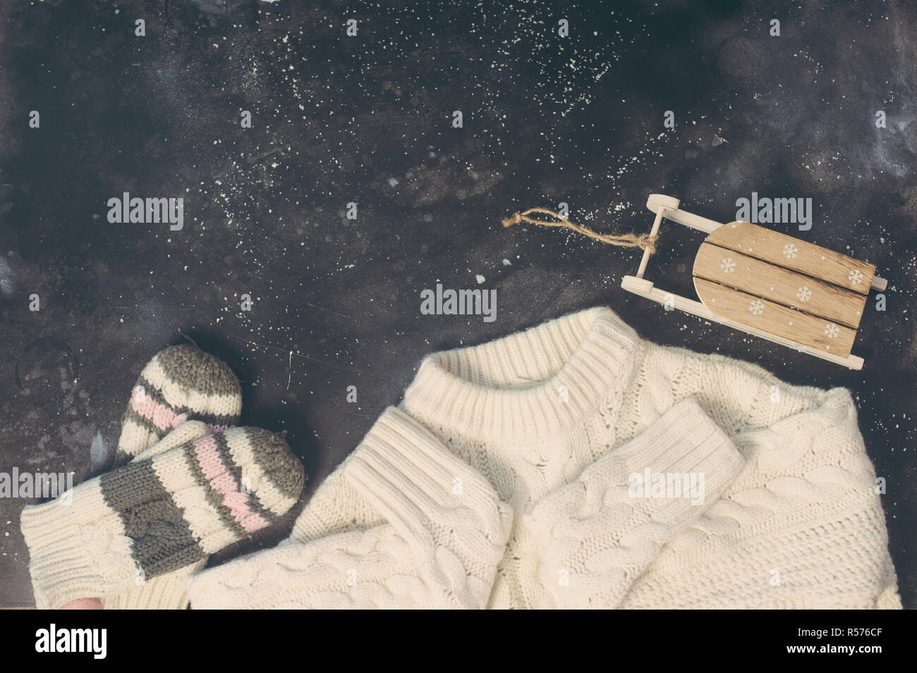 Female winter clothes look on dark concrete. Warm white sweater, knitted mittens and wooden sleigh. Christmas fashion composition top view Stock Photo
