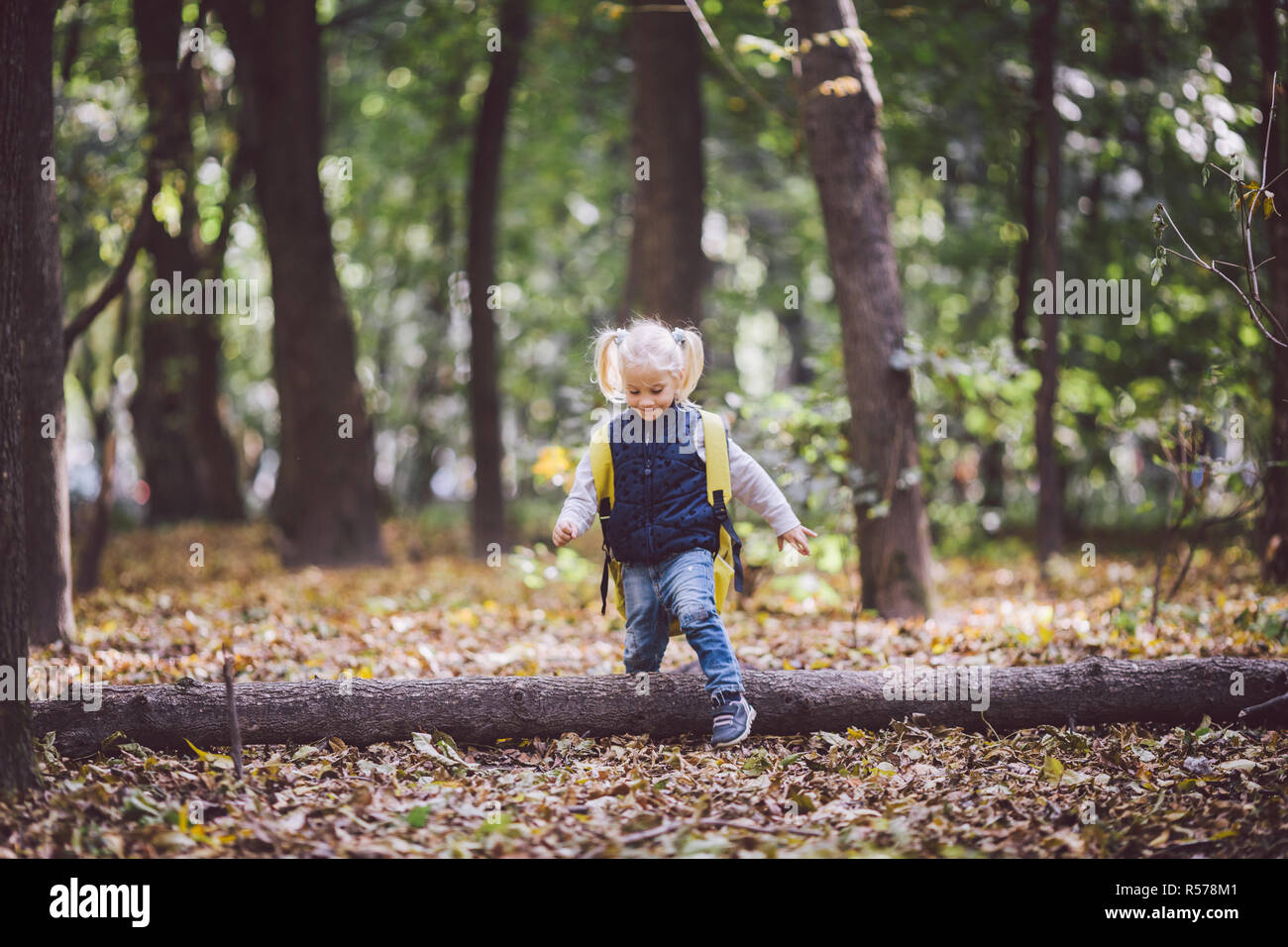 The theme children outdoor activities. Funny little baby Caucasian blond girl walks through forest overcoming obstacles, tree fell, log. Baby hiking big funny backpack in autumn forest park Stock Photo