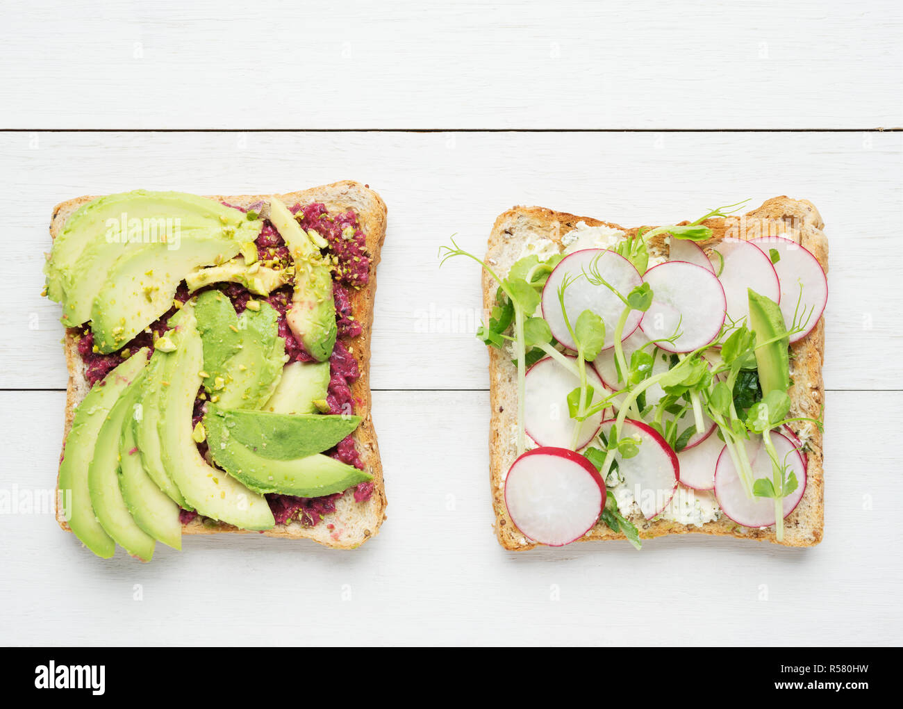 Two avocado toasts with mashed avocado and beetroot,sliced avocado ,radish, snow pea sprouts and goat cheese on white wooden background Stock Photo