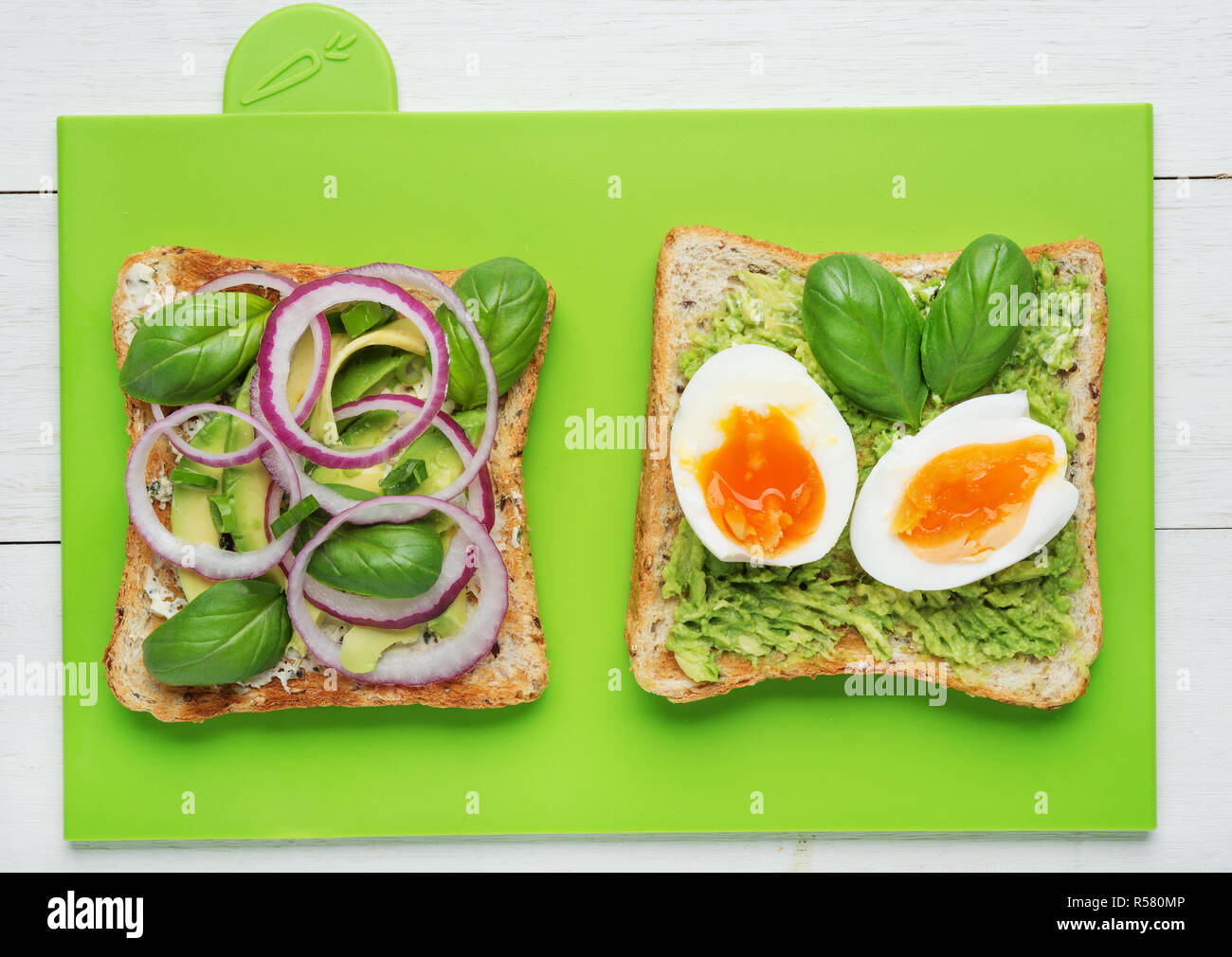 Two avocado toasts with soft boiled eggs,mashed avocado,sliced avocado ,red onion,basil leaves and goat cheese on green background Stock Photo