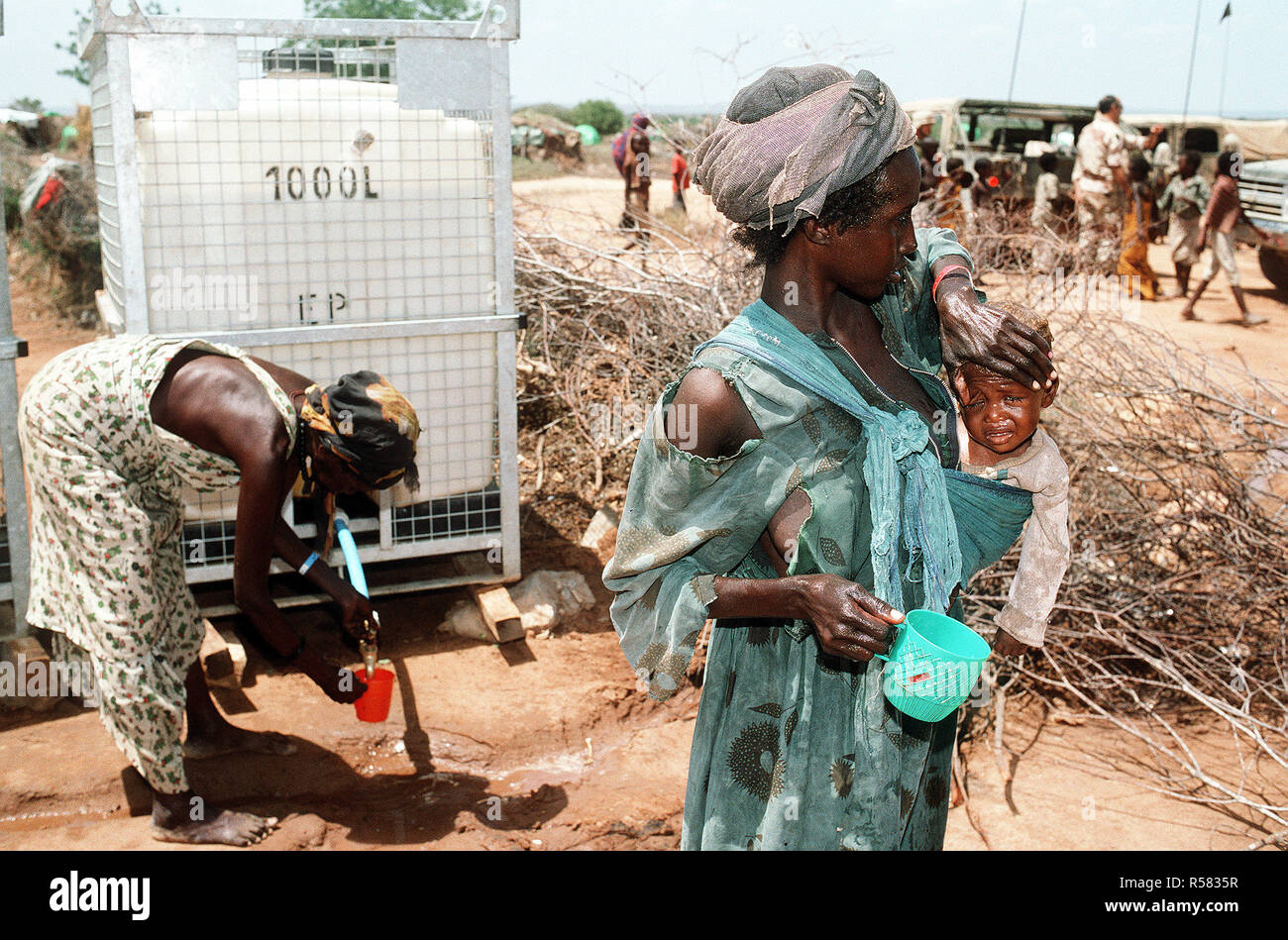A Somali refugee woman holds her child as another woman gets water from a dispenser in the background at an aid station set up during Operation Restore Hope relief efforts. Stock Photo