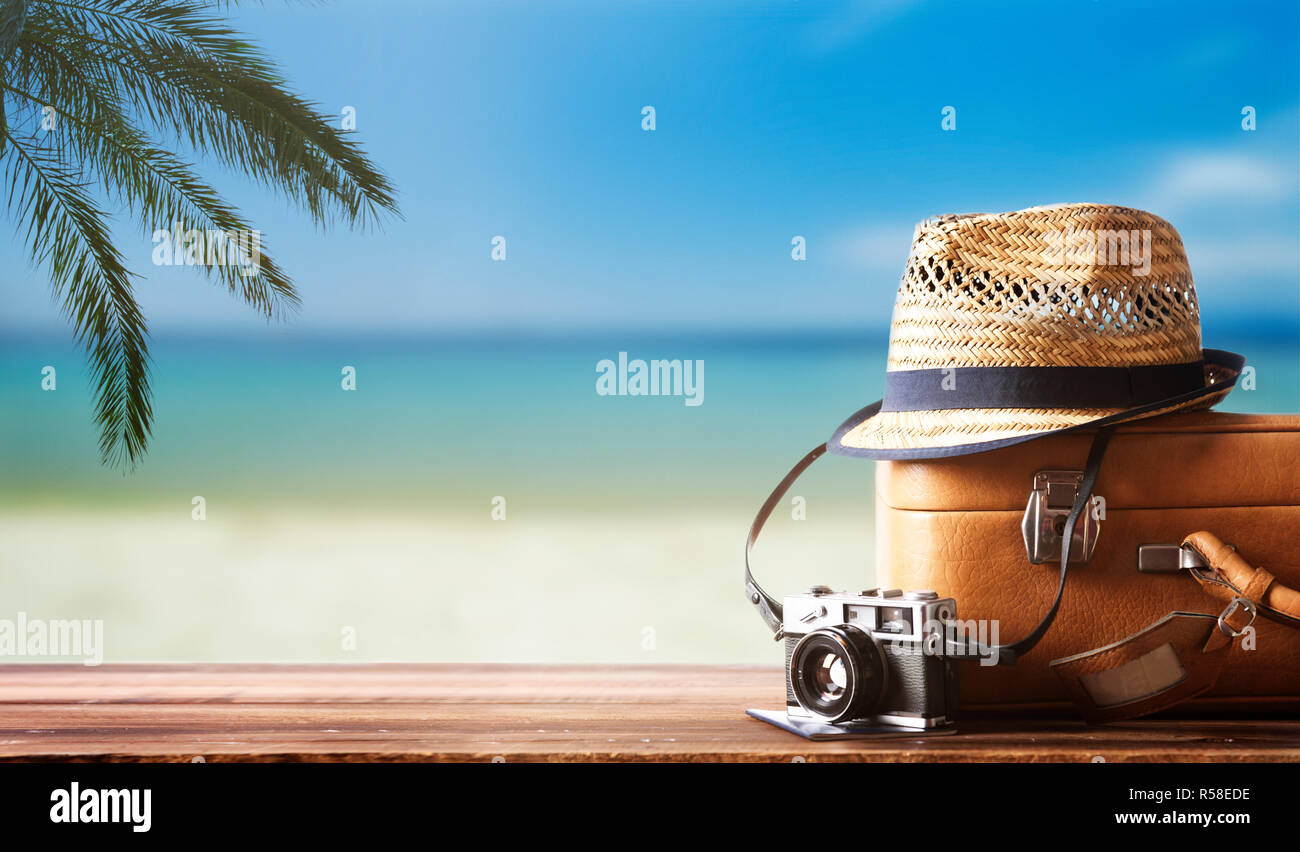 Vintage suitcase, hipster hat, photo camera and passport on wooden dack. Tropical sea, beach and palm three in background. Summer holiday traveling co Stock Photo