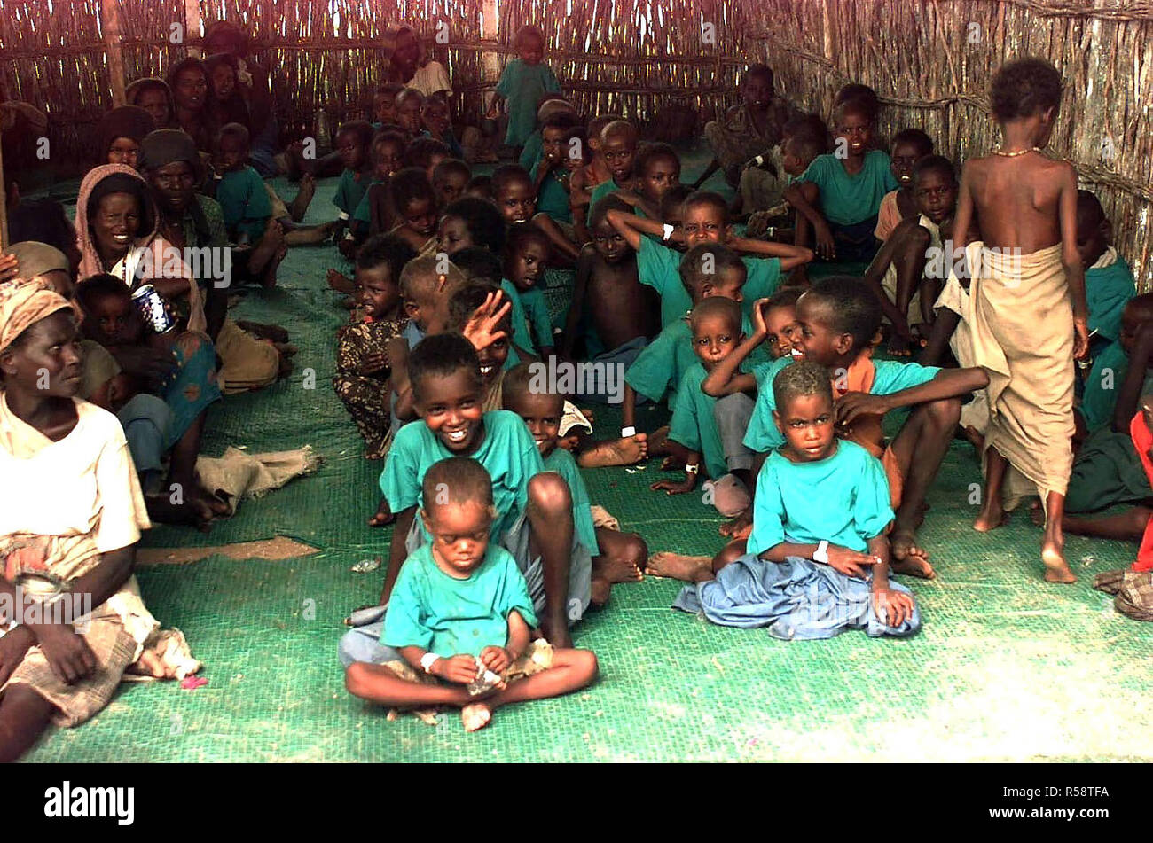 1992 - Straight on shot inside a bamboo hut at several Somali children sitting in rows on a green mat.  Almost all of the children are wearing green t-shirts.  Some older Somali women are seen sitting in a row at the left of the frame. Stock Photo