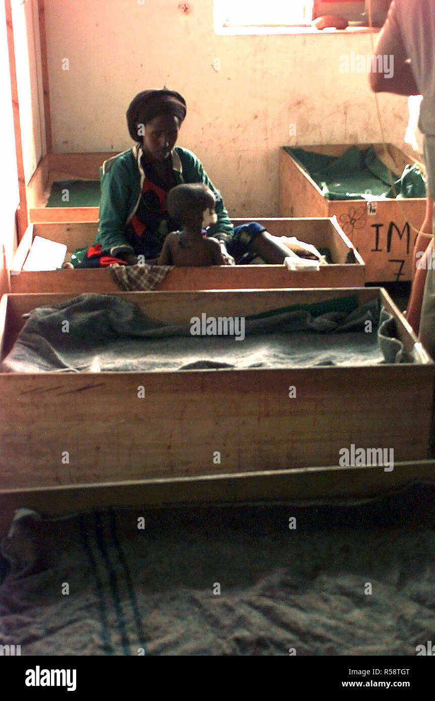 Straight on shot inside a building, at a Somali woman sitting in a wooded box.  A small Somali child sits with its back to the camera.  The box is the child's bed. Stock Photo