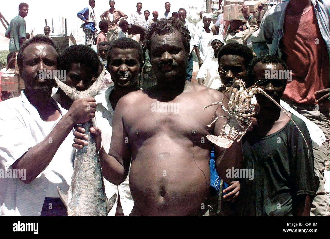 1993 - Two Somali men face the camera and hold up some of the seafood available at a beachside market in Mogadishu.  Four other Somali men stand just behind the first two and face the camera. Stock Photo