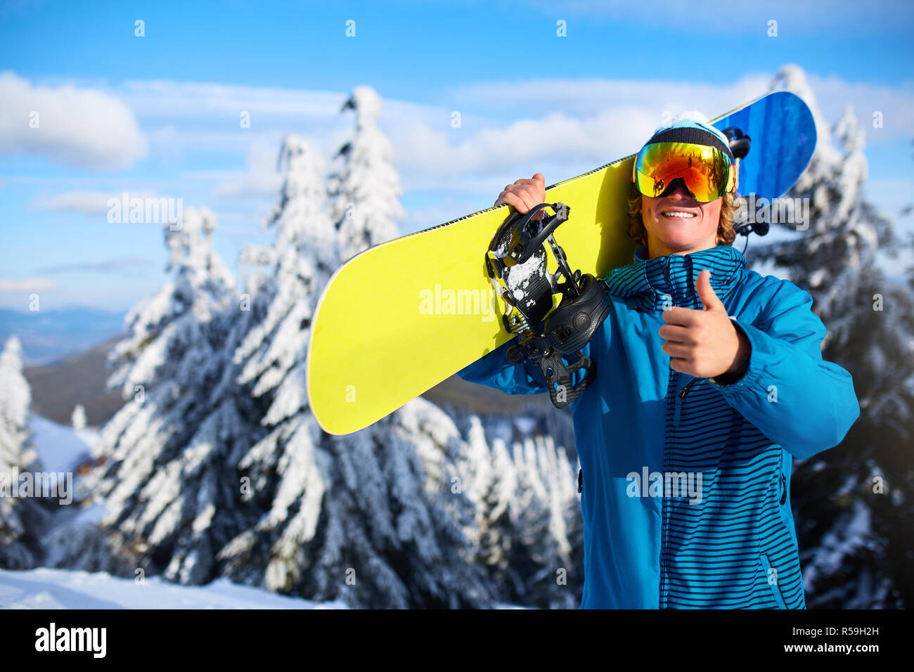 Smiling snowboarder posing carrying snowboard on shoulders at ski resort  near forest before freeride session. Rider showing thumb up sign wearing  polarized goggles. Modern snowboarding equipment Stock Photo - Alamy