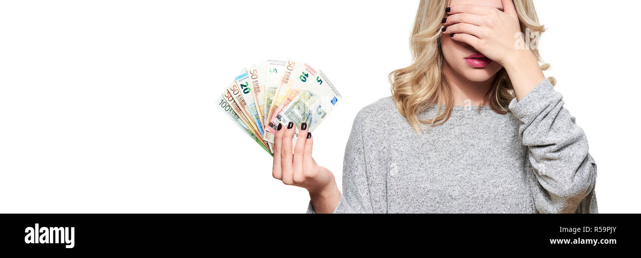 Pretty young woman in grey sweater holding bunch of Euro banknotes, covering her eyes with hand,  isolated on white. Stock Photo