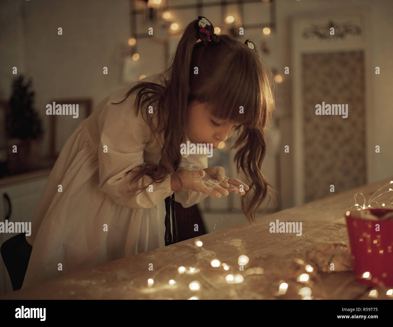 Child girl sprinkles flour on the table and blows on her stained hands on background of garland from light bulbs. Stock Photo