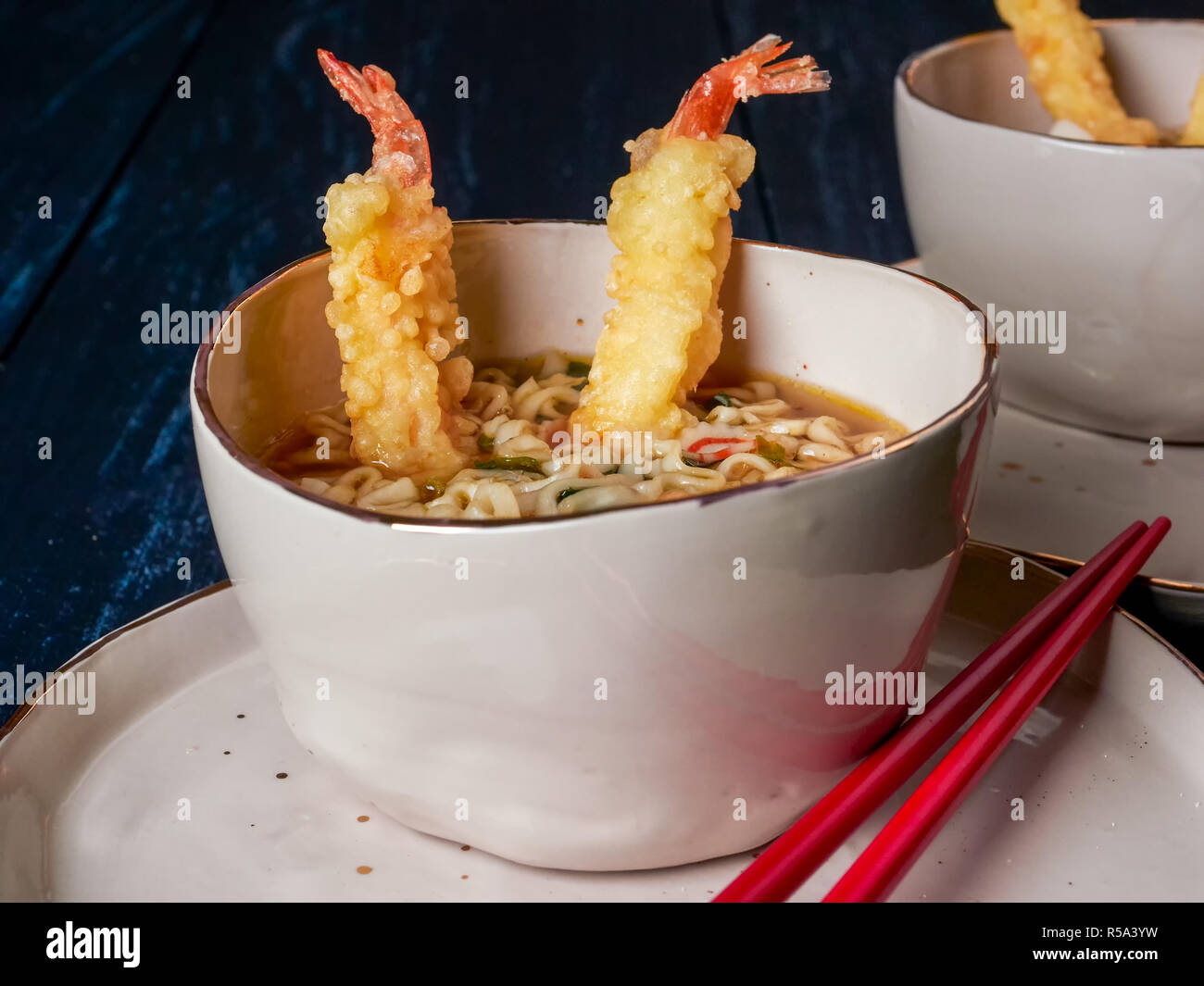 Two bowls of tempura udong noodles, with shrimps temputa, in beige bowls with red chopsticks, on dark blue background Stock Photo