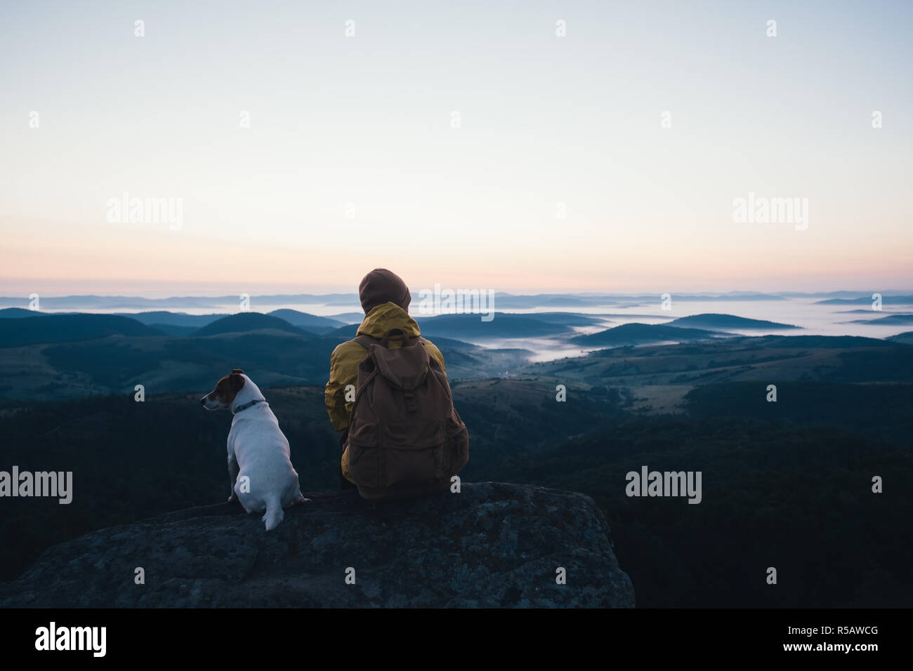 Alone tourist sitting on the edge of the cliff with dog against the backdrop of an incredible mountain landscape. Sunny day and blue sky Stock Photo