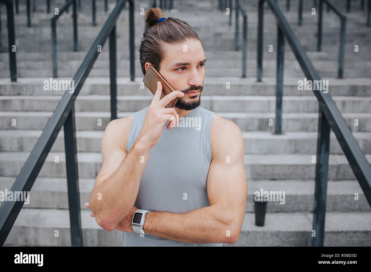 Dark-haired and bearded young man stands and talks on phone. He looks to right. Guy wears handband. There are steps behind him. Guy is alone Stock Photo