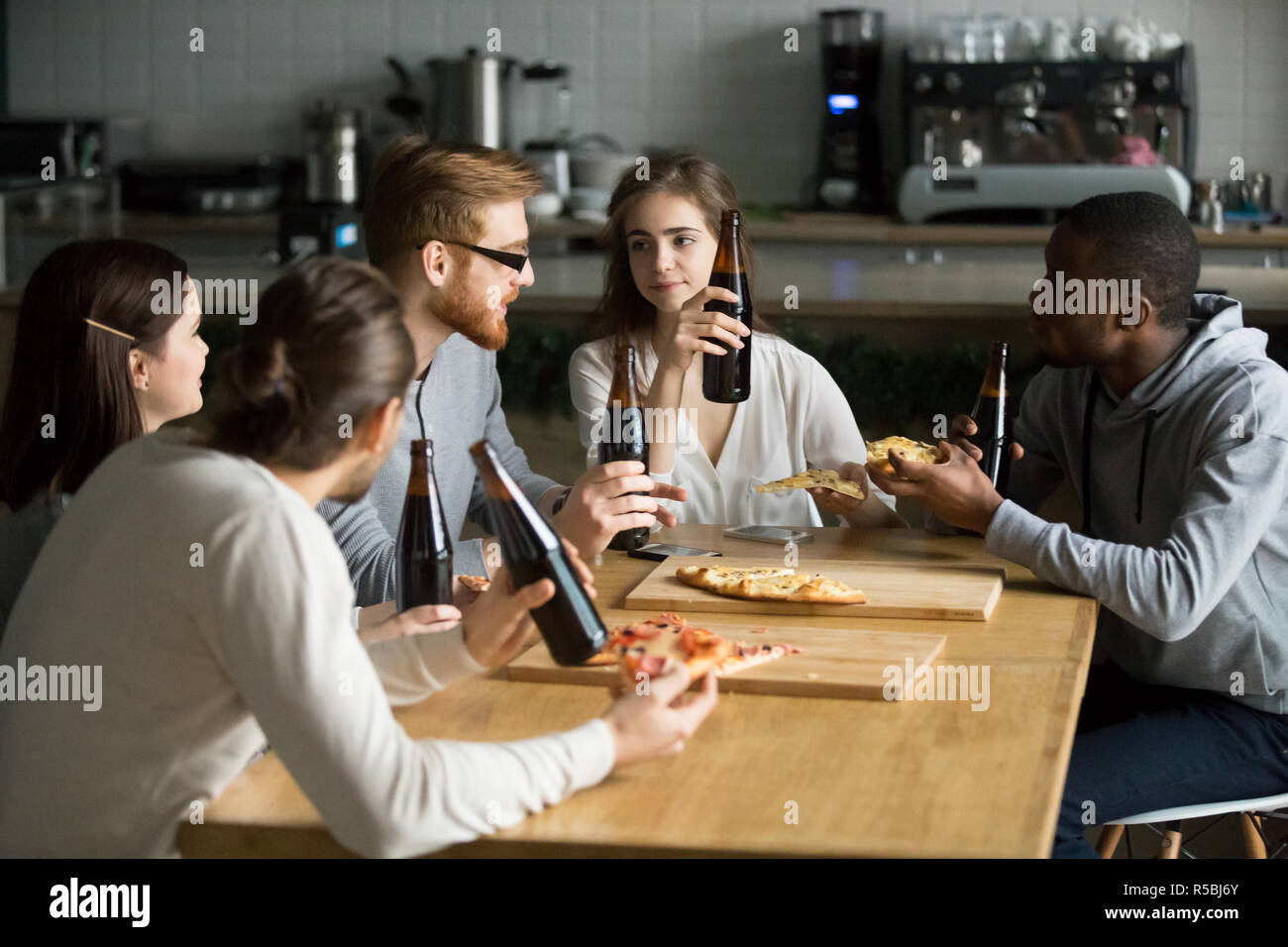 Multiethnic young colleagues meet in Italian restaurant for lunch, diverse friends eat pizza and drink beer, talking hanging out in cafe together, stu Stock Photo