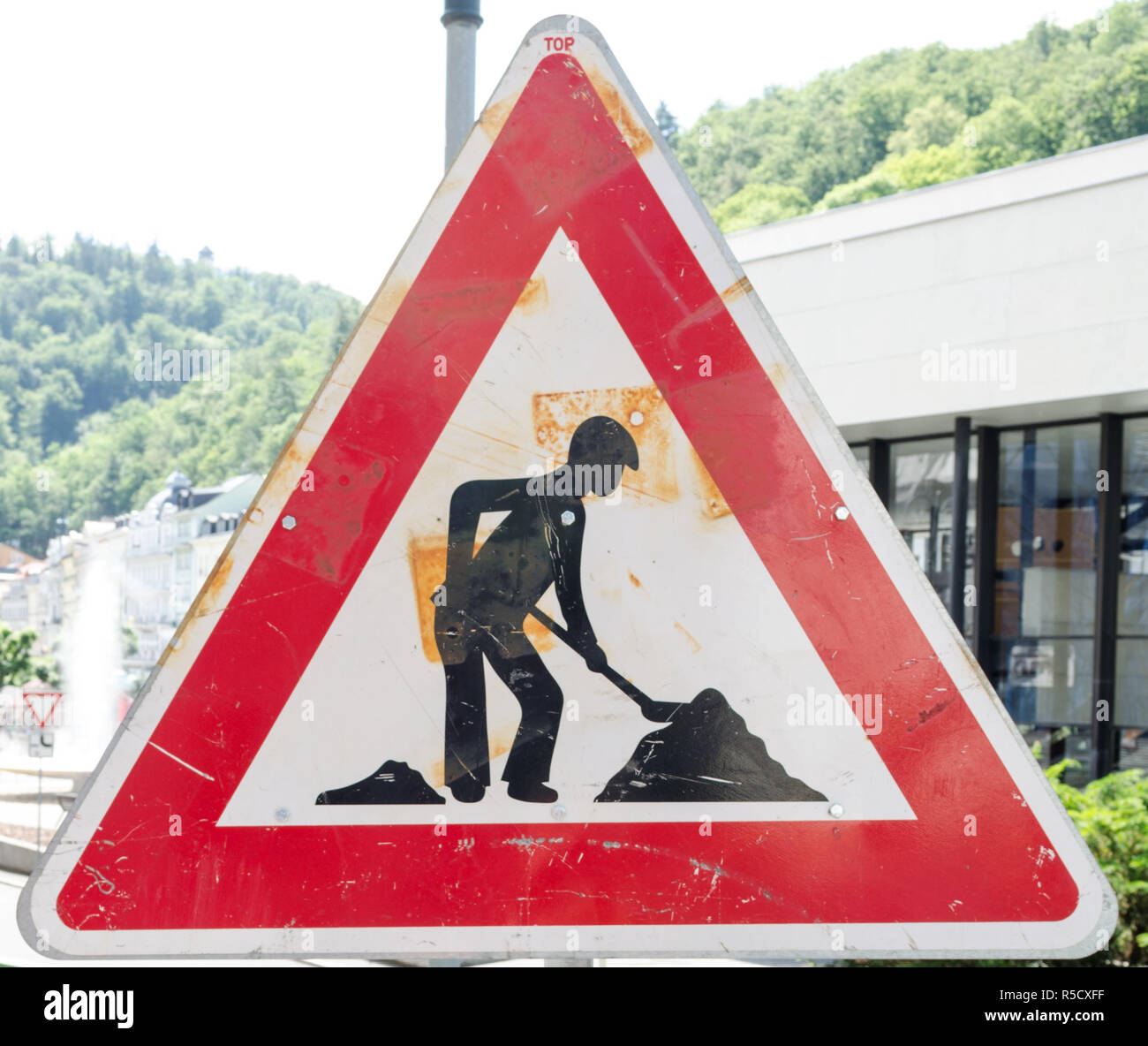 A rusty triangular sign warning of roadworks ahead with a modern city building in the background Stock Photo