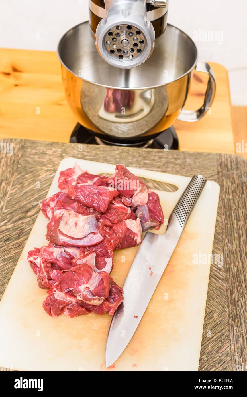Pile of cut raw meat (chuck part) and knife on cutting board with meat grinder in the back. Stock Photo