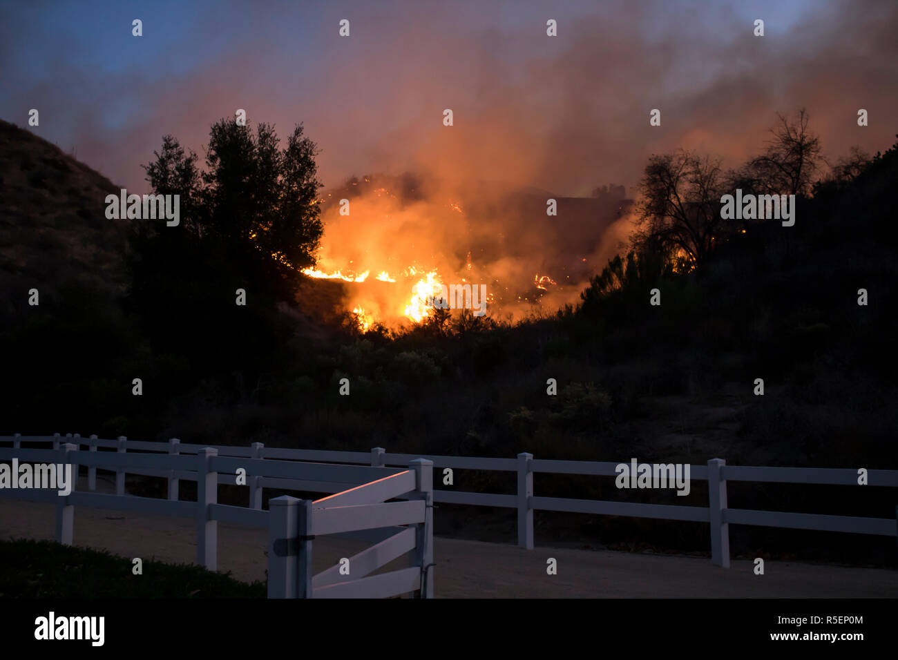 Wildfire flames burn hillside brush at night with dramatic shapes and color in California's Woolsey Fire.  Taken from suburban park with fence in fore. Stock Photo