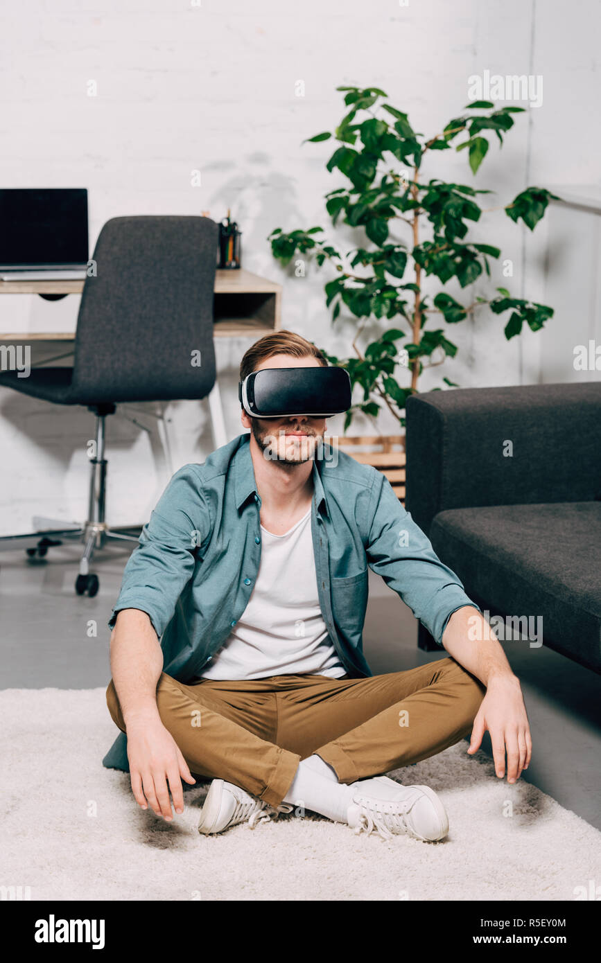young man using virtual reality headset and sitting on floor at home ...
