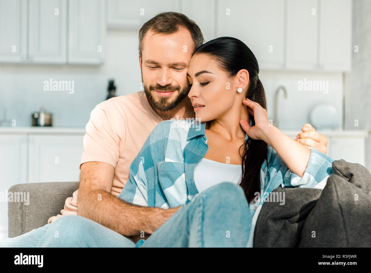 handsome husband and beautiful wife sitting and embracing on couch at