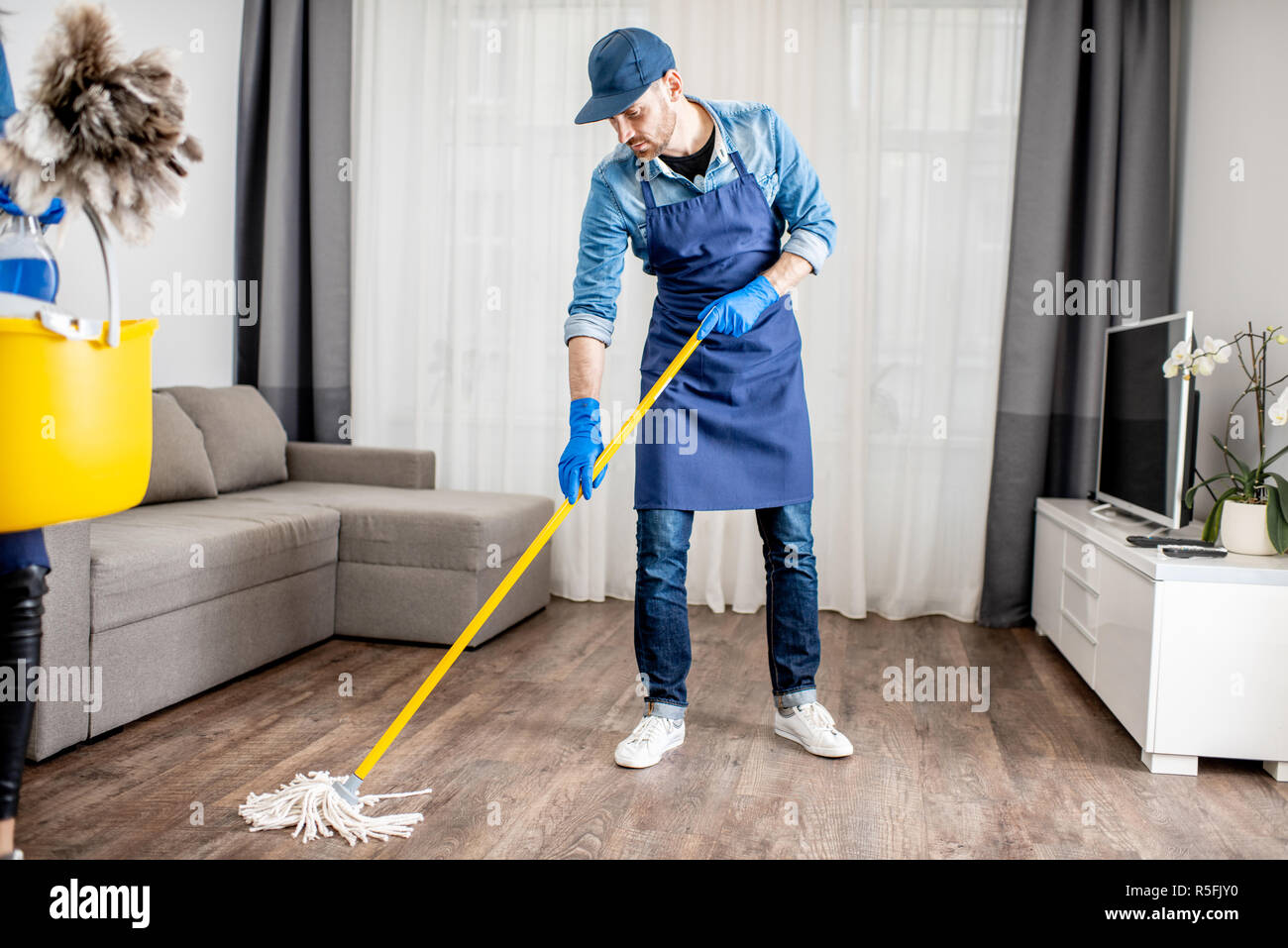 Man as a professional cleaner dressed in the blue uniform washing floor with mopping stick in the apartment Stock Photo