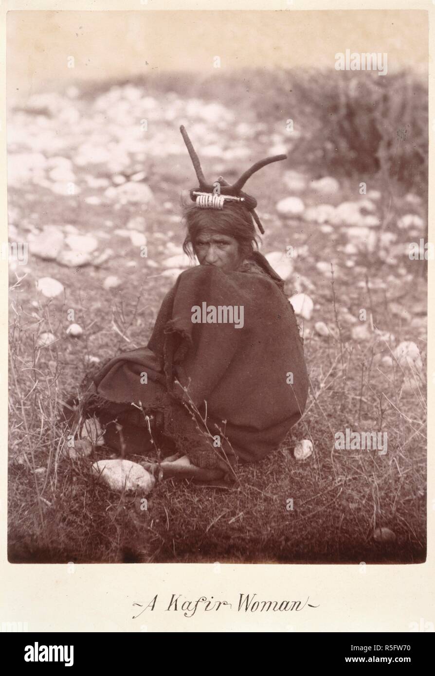 A Kafir woman. Full-length portrait of a seated woman. The photograph shows the characteristic headdress of married Bashgal women: 'Here, for the first time, Bashgal women were seen in their own country by any European, and the novel experience was not pleasing. They had hard, forbidding faces, and were very dirty. Their only dress was a long goat's-hair gown, reaching from the neck to below the knee, and they, like the men, wore ankle boots of soft brown leather. The married women wore a head-dress, but only out of doors. Their head-dress is peculiar to the Bashgal Kafirs. It consists of a ca Stock Photo