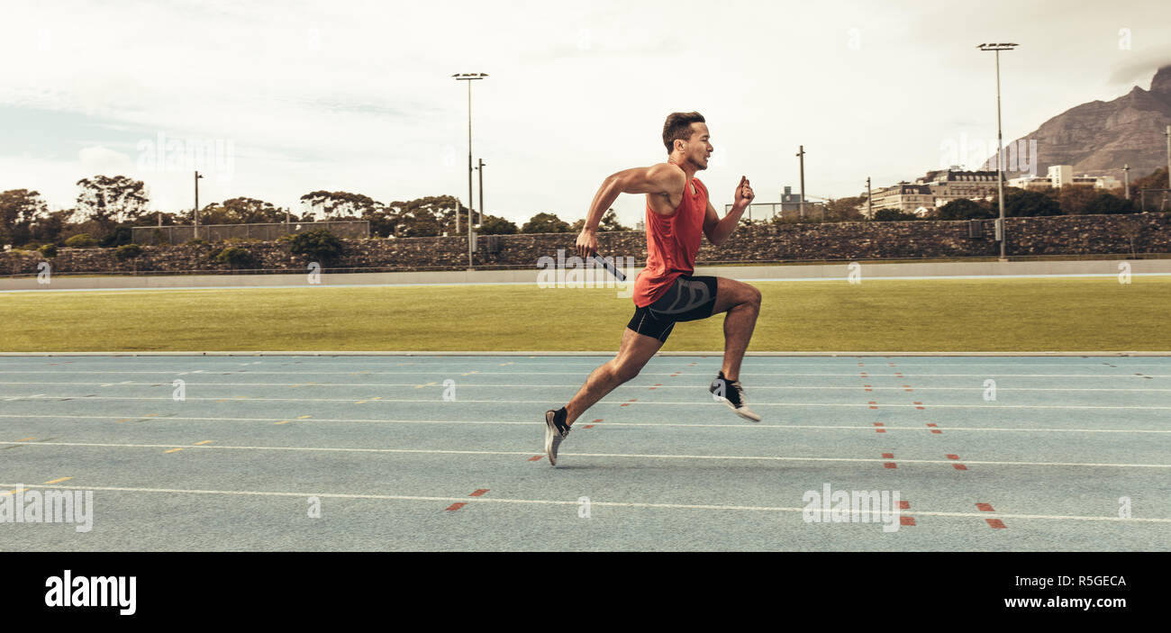 Side view of a male athlete sprinting on a running track in a track and field stadium holding a baton. Male runner training on a running track. Stock Photo