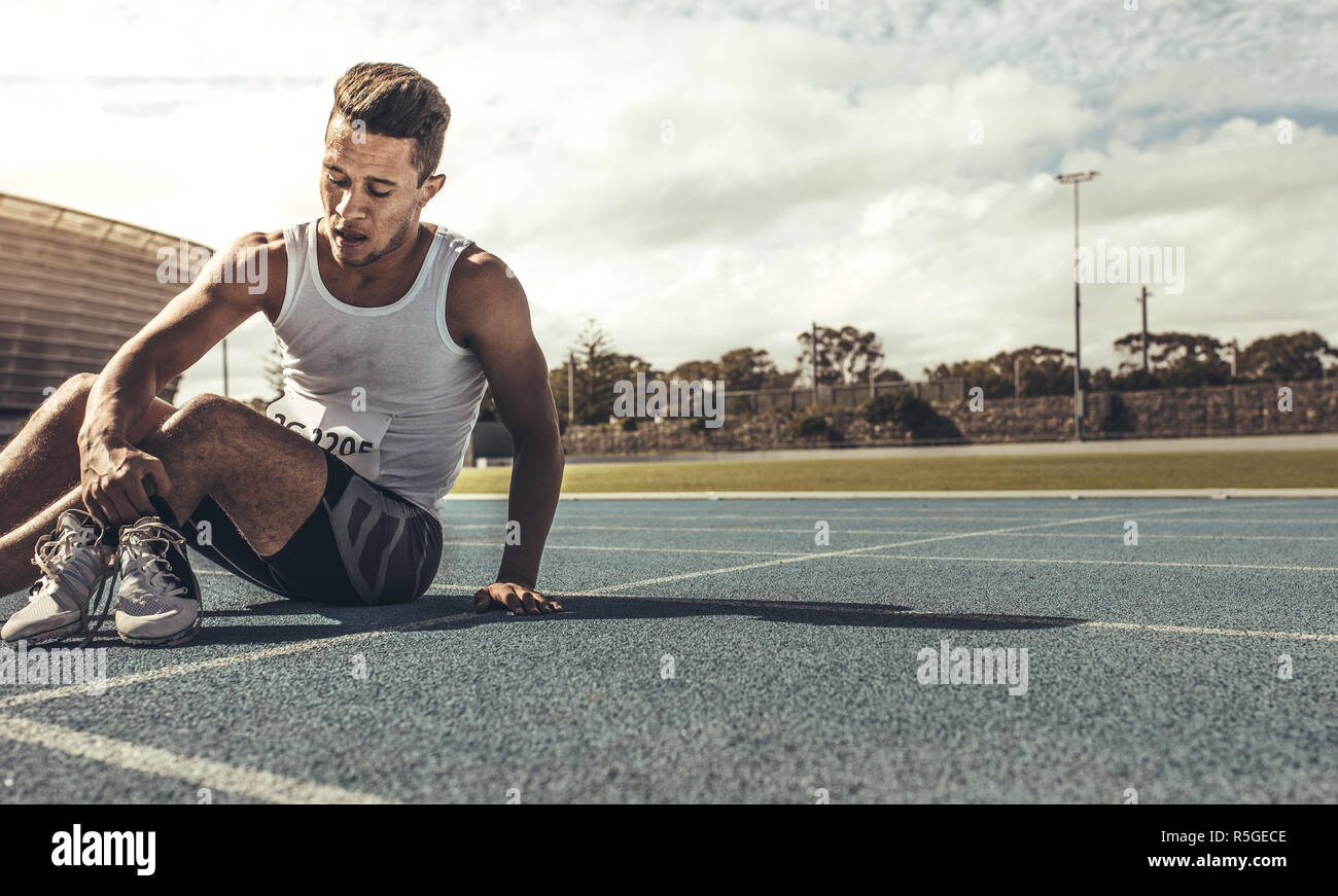 Athlete taking off his sprinting shoes after the race sitting on the track. Tired athlete relaxing on field after the race holding his shoes. Stock Photo