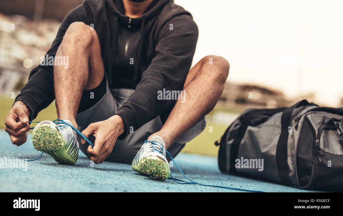 Athlete wearing sports shoes getting ready for training. Cropped shot of a runner sitting on a running track tying shoe lace with a bag by his side. Stock Photo