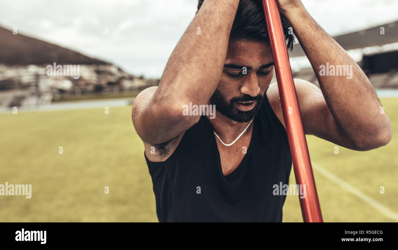 Tired athlete standing in a track and field ground holding a javelin. Athlete relaxing during his javelin training standing with his eyes closed holdi Stock Photo