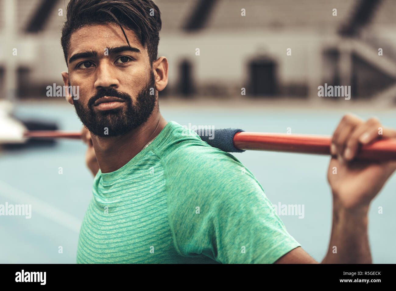 Athlete standing in a stadium holding a javelin on his shoulders. Man training in javelin throw standing in a stadium looking away. Stock Photo