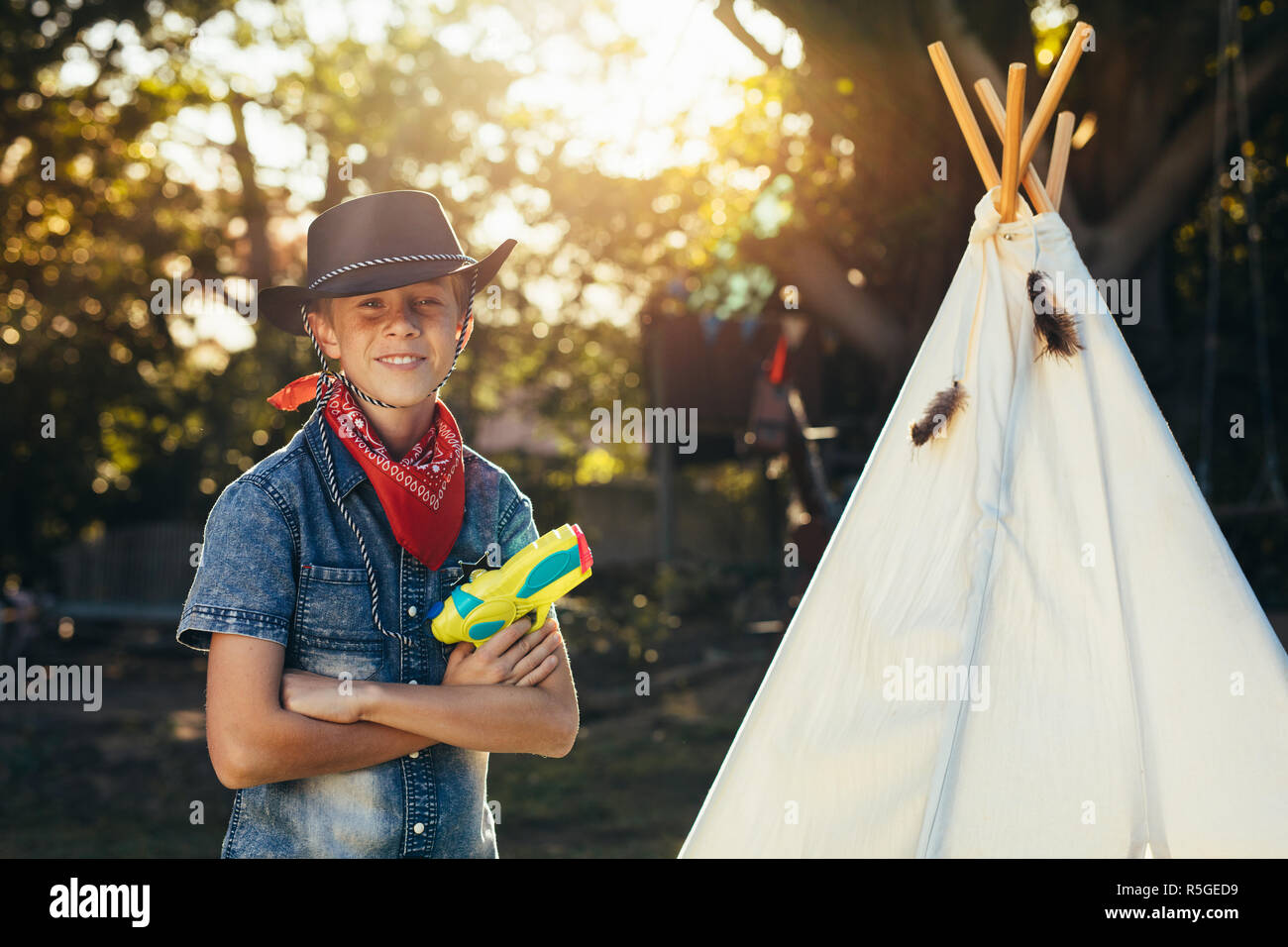 Little cowboy standing outdoors by a toy hut in backyard with water pistol. Young boy wearing cowboy hat with water squirt gun. Stock Photo