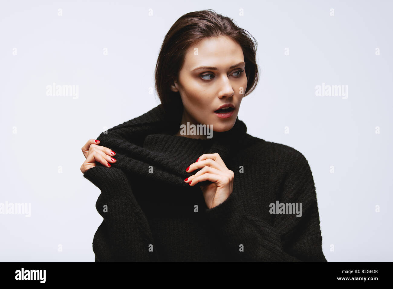 Attractive woman pulling the collar of her turtleneck sweater on white background. Caucasian female model with surprised look. Stock Photo