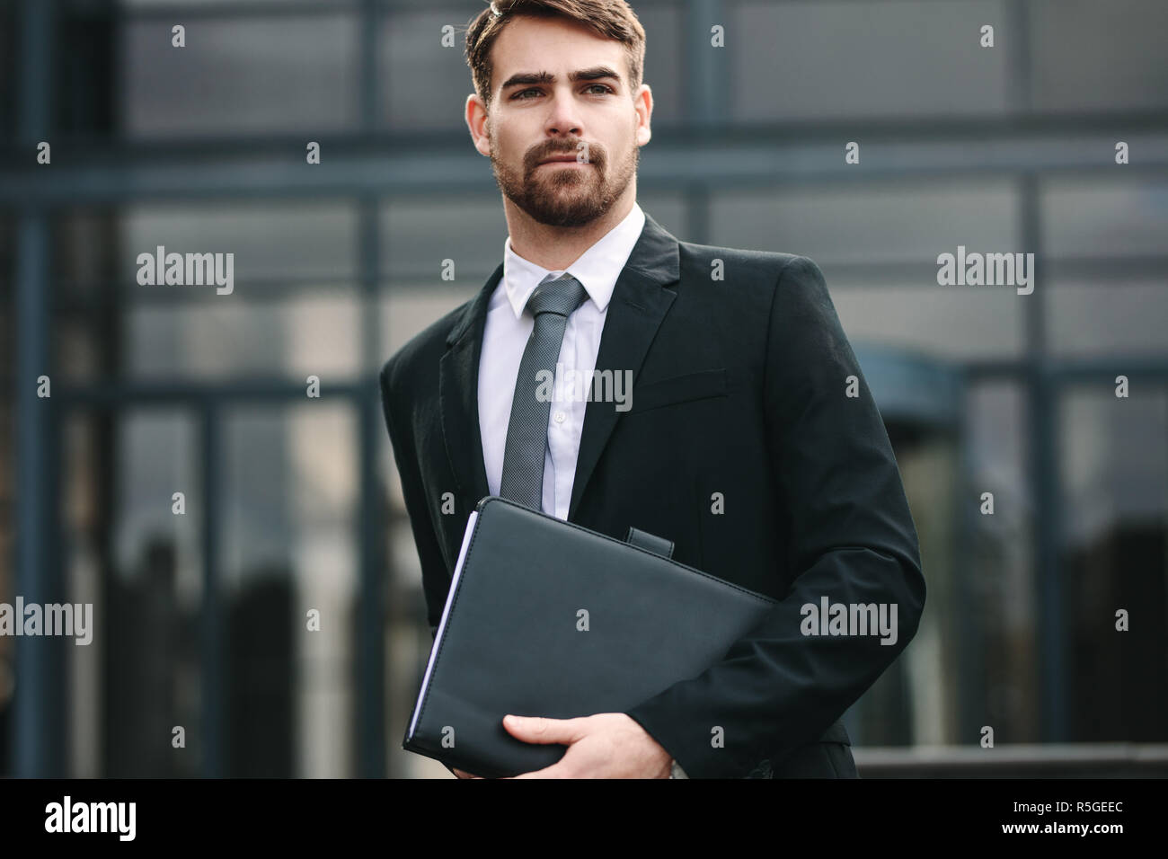Portrait of handsome businessman with a folder walking outdoors. Man in businesswear holding a file and looking away. Stock Photo