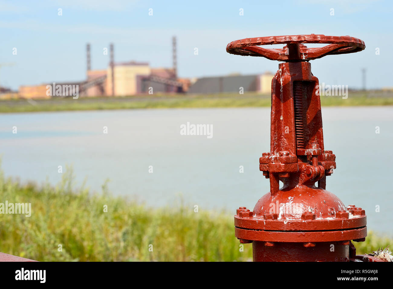 The valve from the pipeline against the background of the factory and an artificial lake Stock Photo