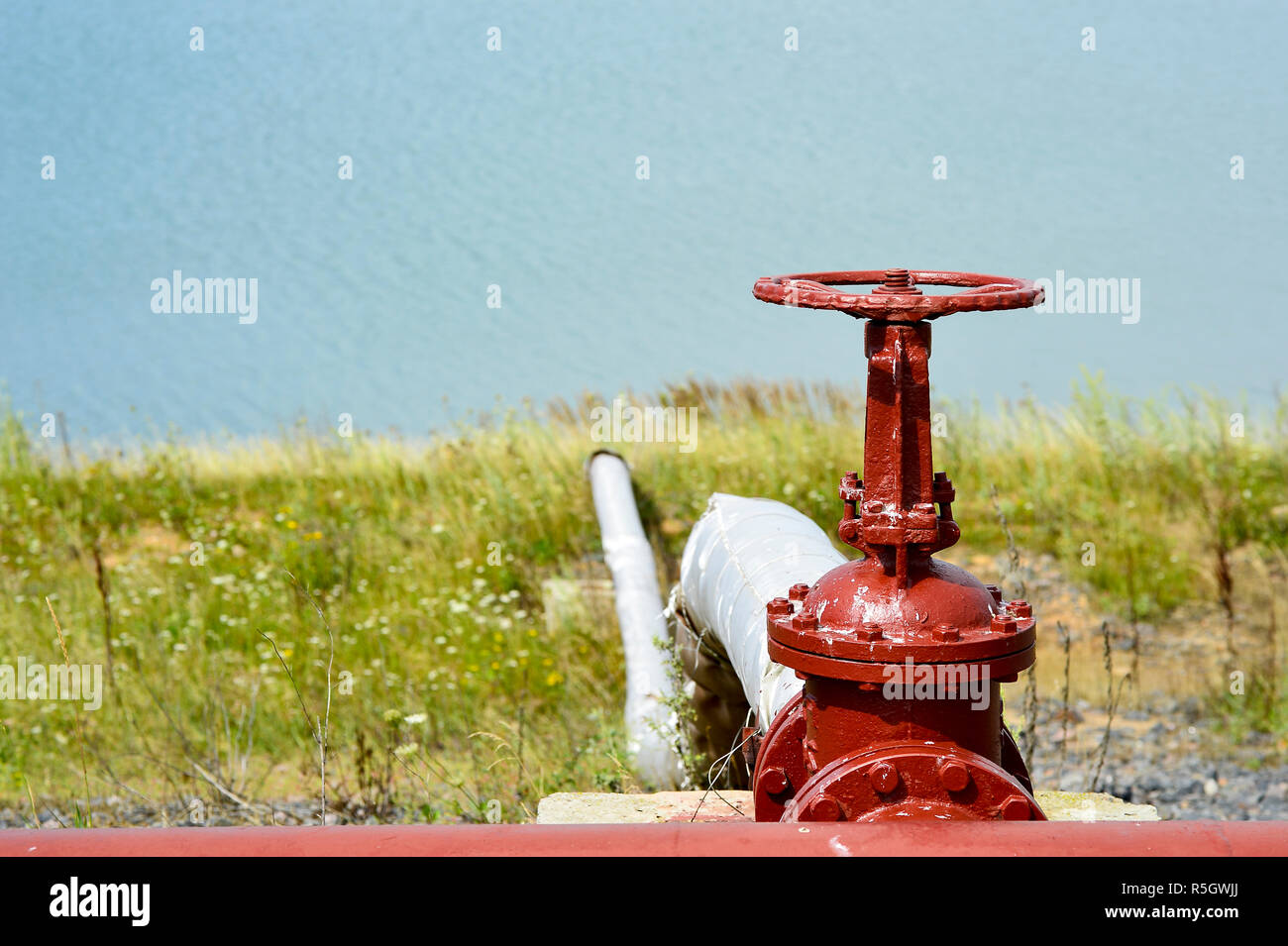 The valve from the pipeline against the background of the factory and an artificial lake Stock Photo