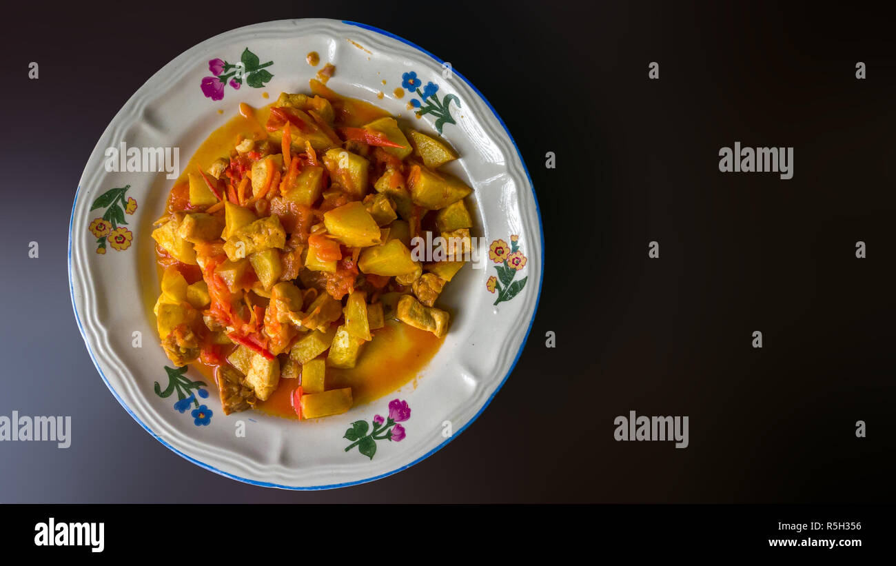 Authentic Indonesian food from family street restaurant. Mixed tofu potato and tempe cooked in tomato and carrot sauce. Plate isolated on plain dark b Stock Photo