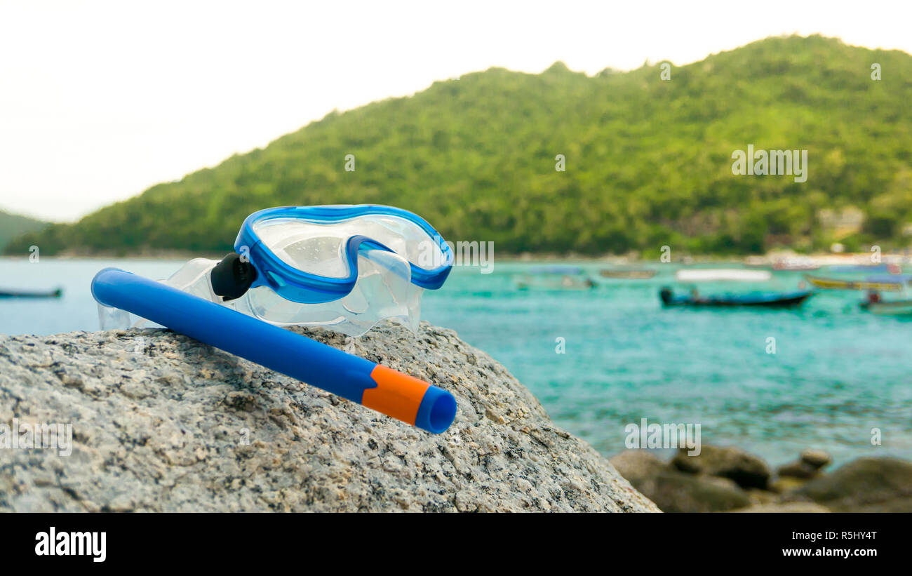 Snorkeling equipment: snorkel and diving google on the stone. Tropical island, sea, beach and boats in background. Stock Photo