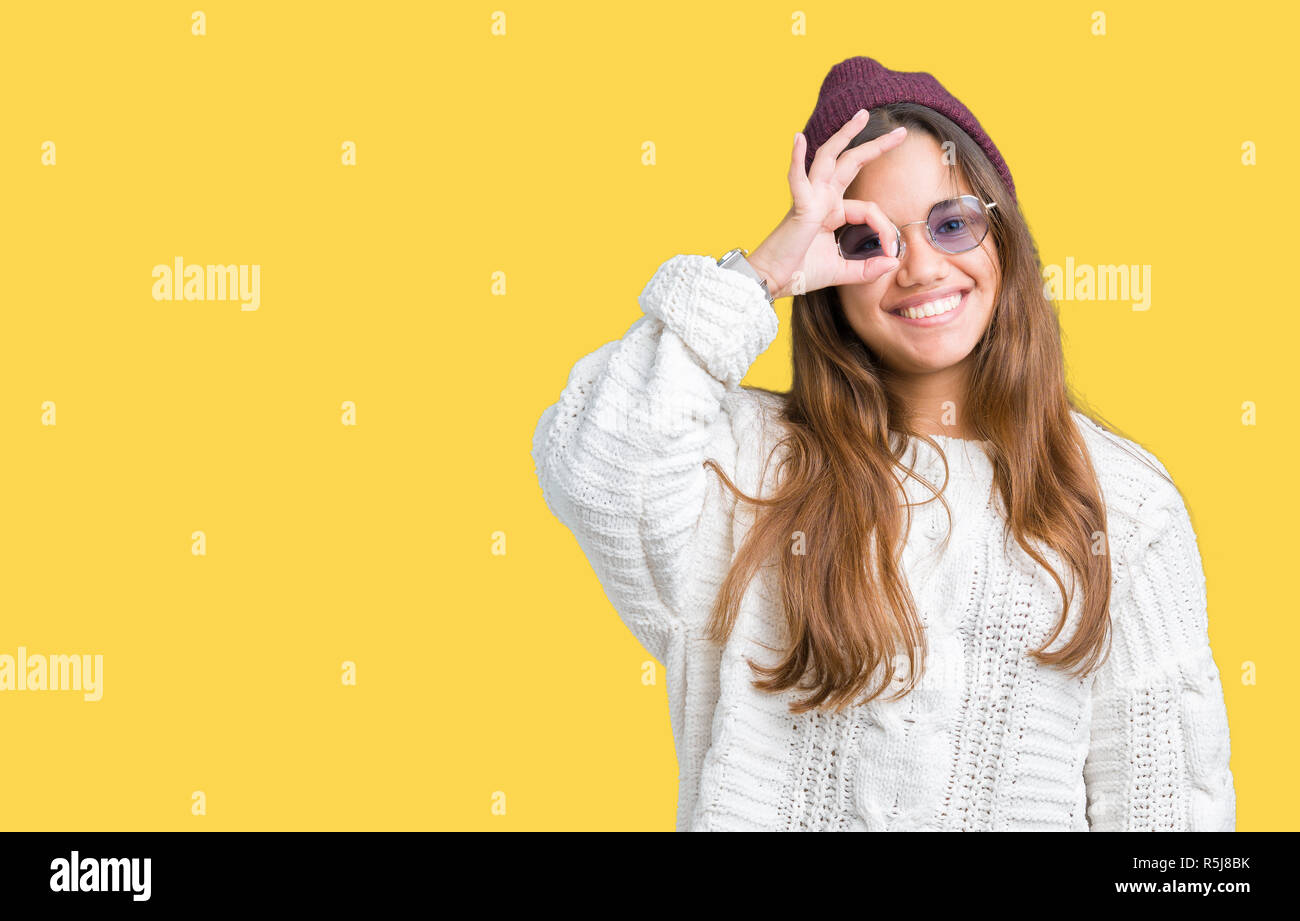 Young beautiful brunette hipster woman wearing sunglasses over isolated background doing ok gesture with hand smiling, eye looking through fingers wit Stock Photo