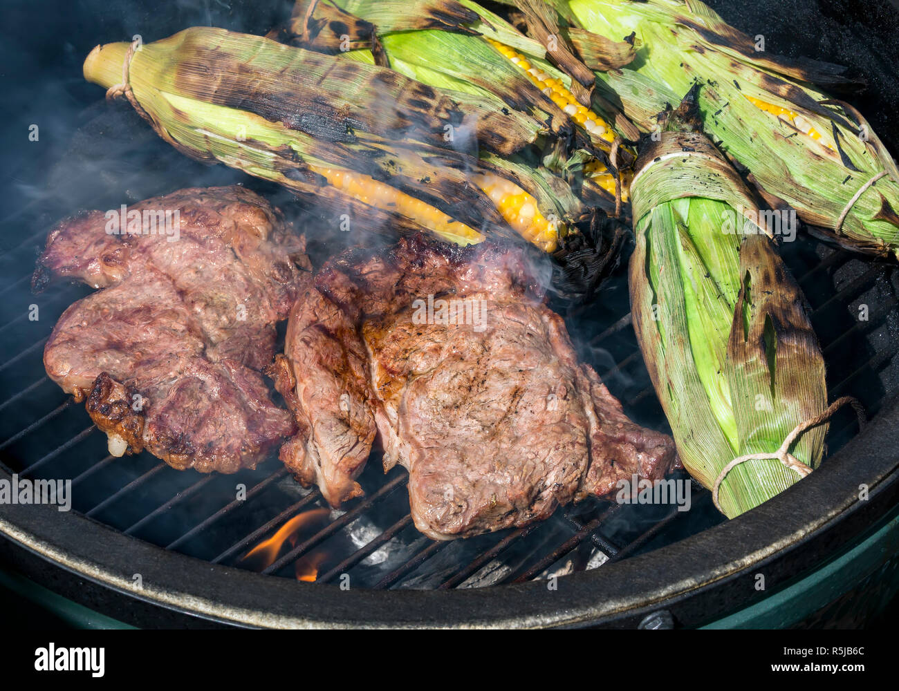 Two rib eye steaks and ears of sweet corn in the husks cook on a smokey