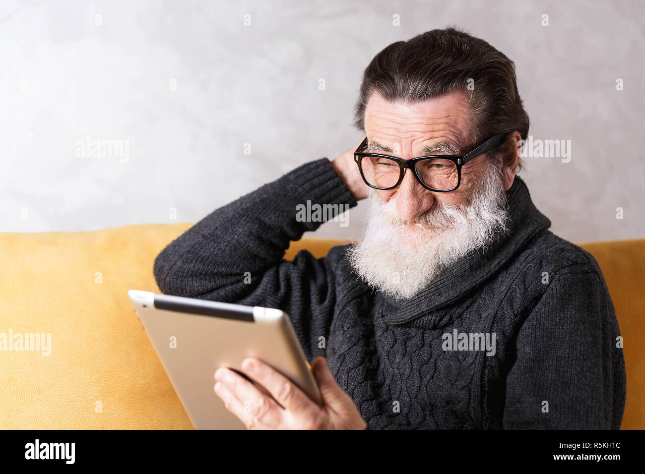 Senior bearded man in glasses wearing grey pullover sitting on a yellow sofa in his light living room and looking doubtfully on the digital tablet Stock Photo