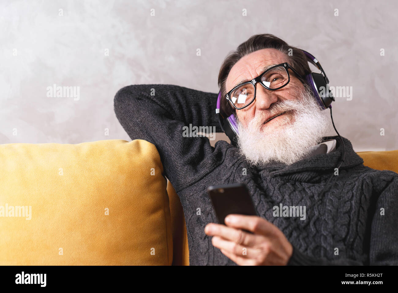 Senior cheerful bearded man in glasses wearing grey pullover relaxing with the smartphone while sitting in headphones on a yellow sofa in his light living room, modern technology, communication concept Stock Photo