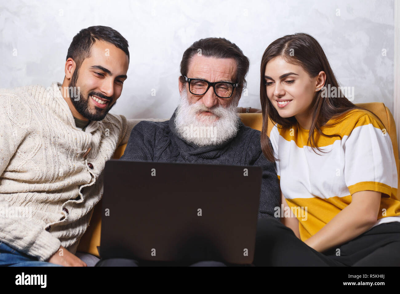 Senior bearded father in glasses sitting with his children on the yellow sofa in the light living room and watching movie, modern technology, communication concept Stock Photo