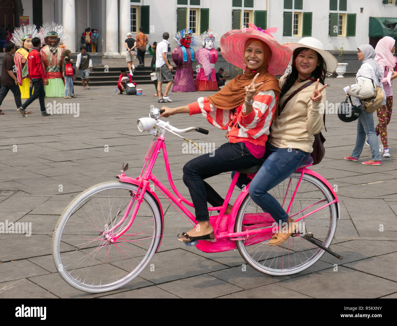 Horizontal photo of two smiling, happy, young Indonesian girls riding a pink bike with white tires wearing big hats in Jakarta Old Town Square. Stock Photo