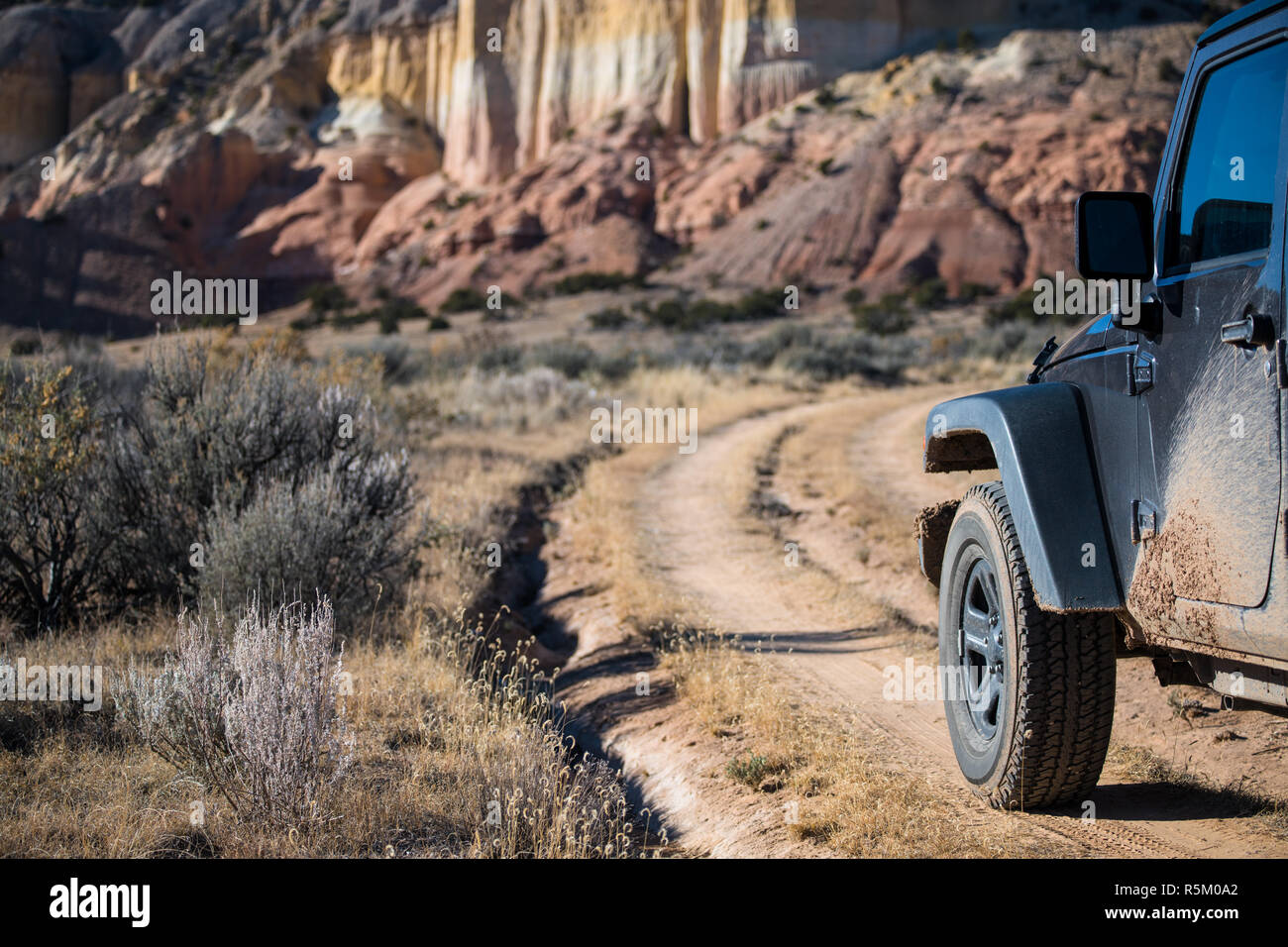 Muddy four-wheel drive vehicle on curving dirt road heading toward colorful cliffs in the high desert landscape in the Rio Chama canyon near Santa Fe, Stock Photo
