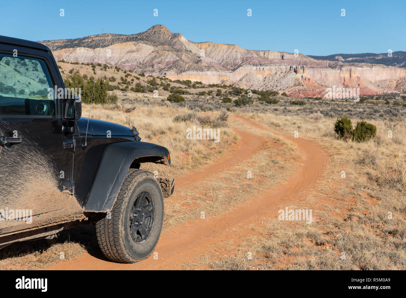 Muddy four-wheel drive vehicle on curving dirt road heading toward a colorful high desert peak in Ghost Ranch near Santa Fe, New Mexico Stock Photo
