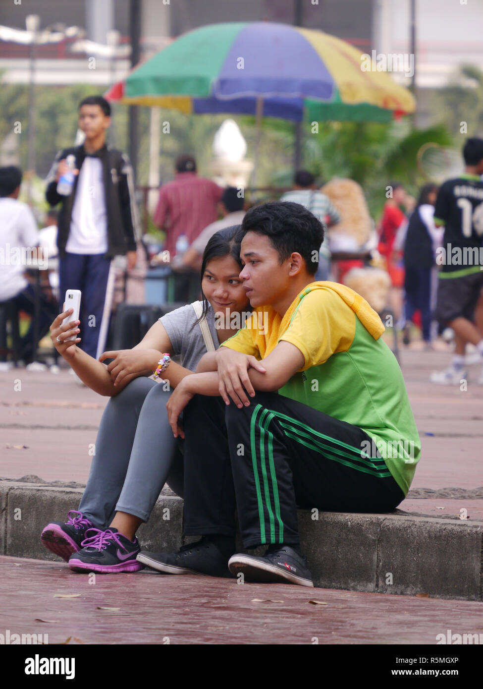 Horizontal photo of content, young Indonesian teenage girl resting her head on boyfriend's shoulder looking at cell phone of selfies taken that day. Stock Photo