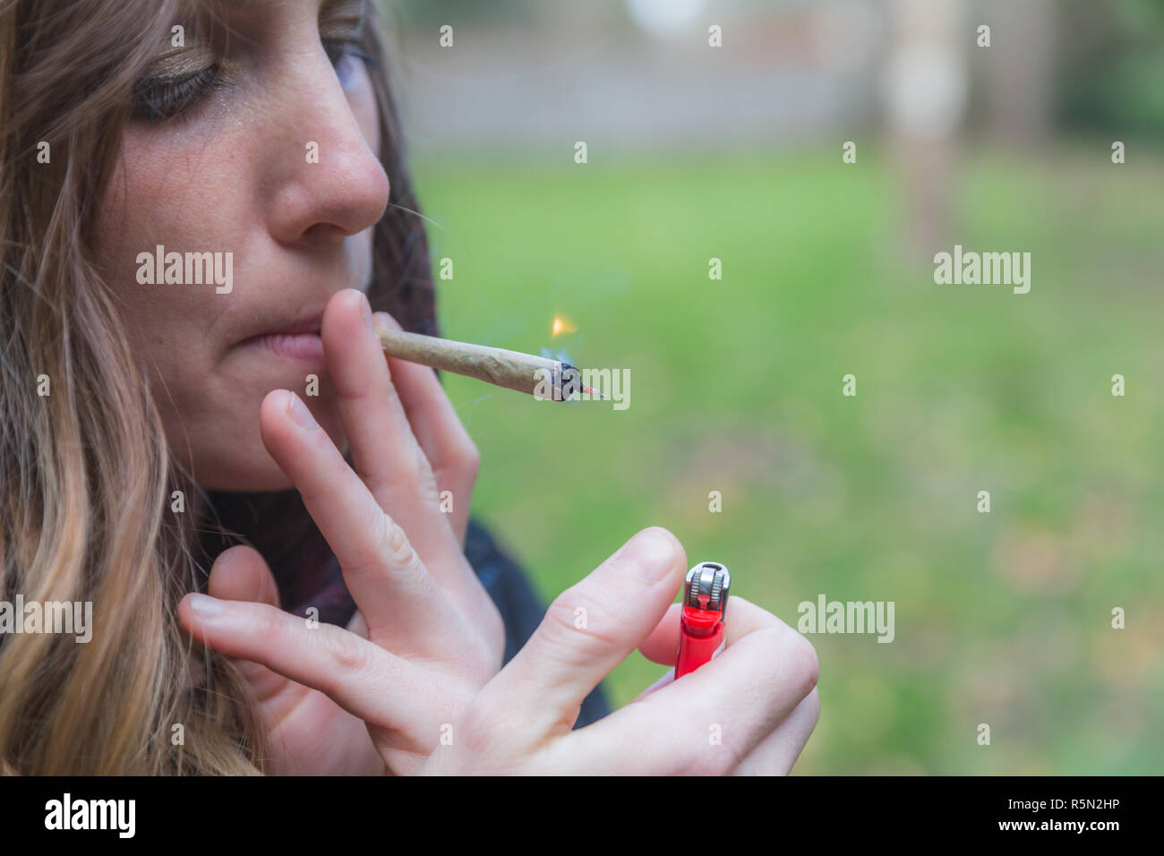 Young woman with blonde hair lighting a rolled joint and smoking it outdoors. Stock Photo