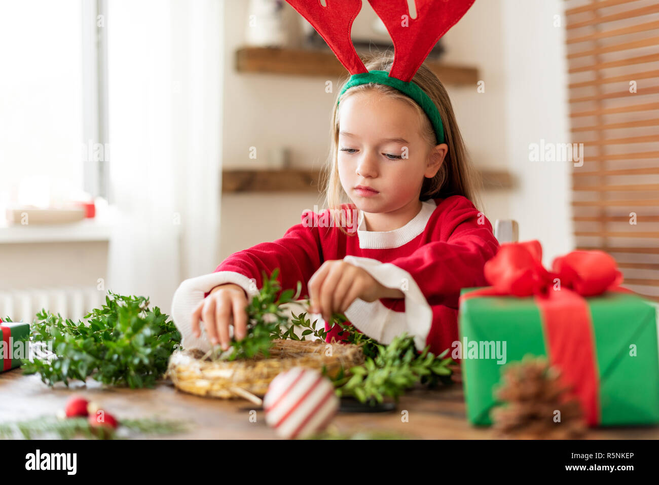 Cute preschooler girl dressed in reindeer costume wearing reindeer antlers making christmas wreath in living room. Christmas decoration family fun con Stock Photo