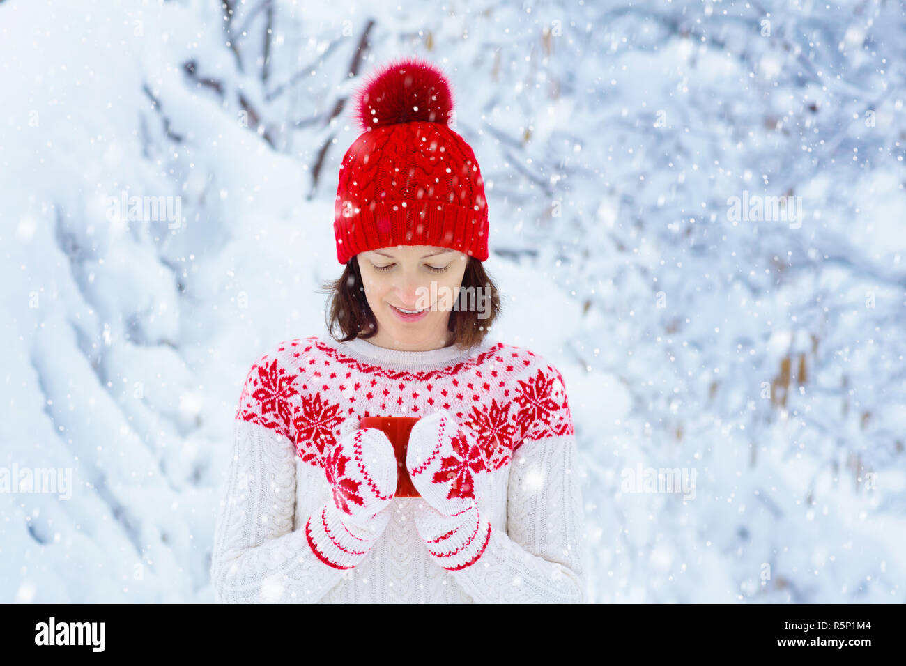Woman drinking hot chocolate in Christmas morning in snowy garden. Girl in knitted Nordic sweater, hat and mittens holding cup with cocoa. Snow and wi Stock Photo