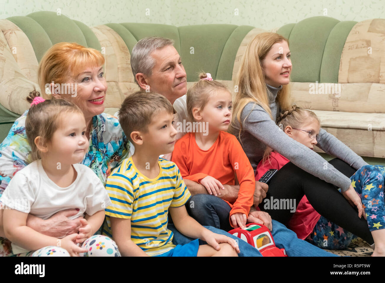 Grandfather and grandmother with their daughter and grandchildren watching television sitting on the floor by the sofa Stock Photo